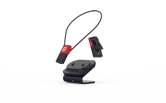 The image shows three main components: 1) A small black and red lavalier microphone  connected to a dark lanyard, 2) A clip-on attachment for the microphone for attaching to clothing, and 3) A black double-slot charging dock. The microphone features the Catchbox logo in white on its red jacket. 