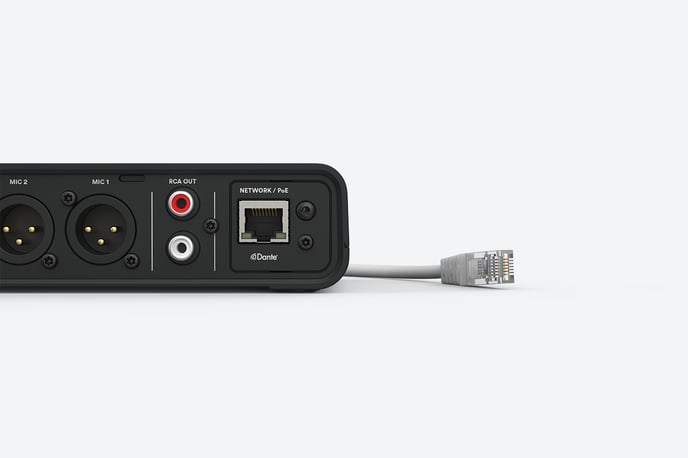 A black Hub receiver is shown with several ports visible on its back panel. From left to right, there are two XLR ports labeled "MIC 2" and "MIC 1," two RCA output ports (one red and one white) labeled "RCA OUT," and an Ethernet port labeled "NETWORK/PoE" with a Dante logo. An Ethernet cable is partially plugged in, lying next to the receiver.